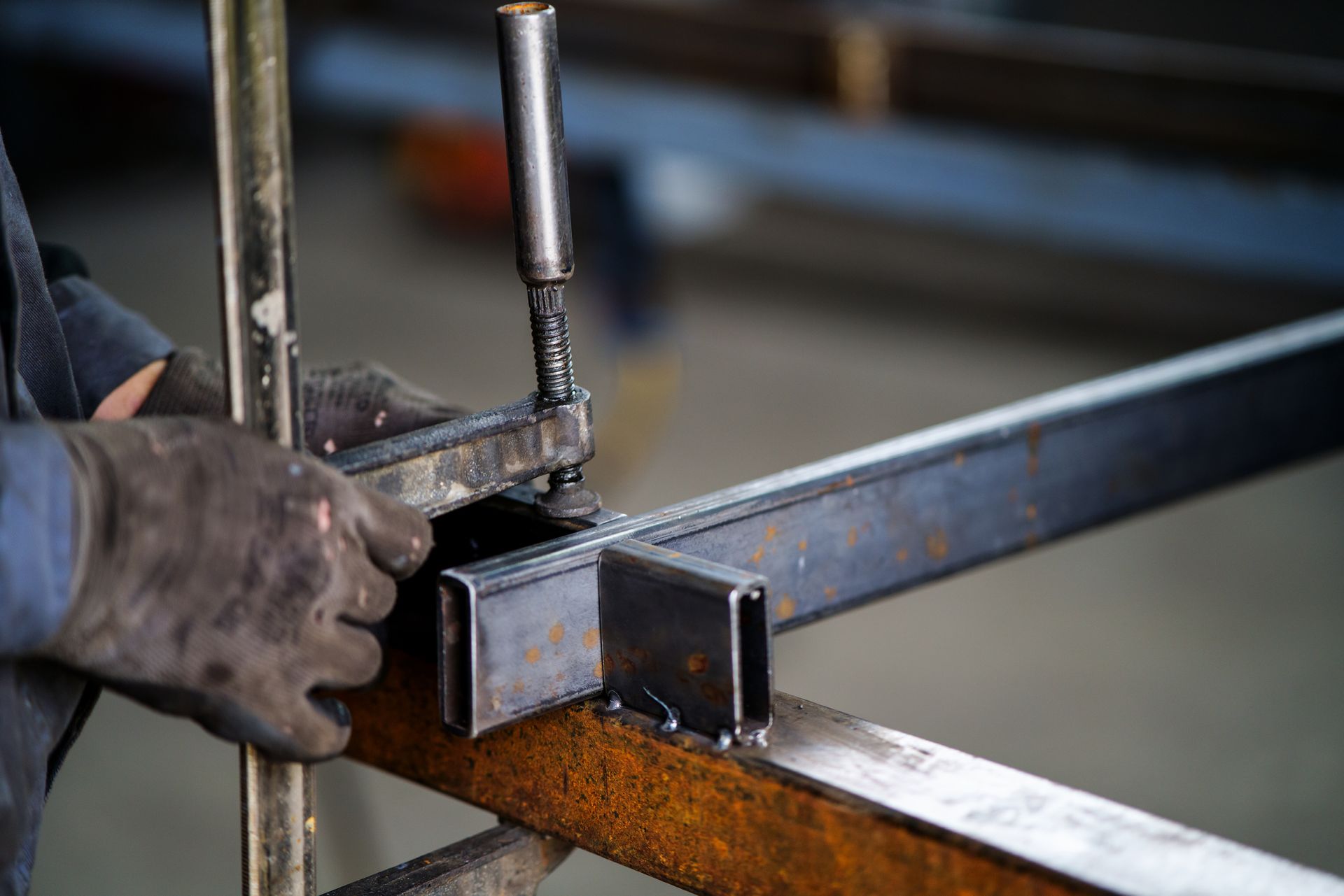 A worker welds metal beams together in a workshop, wearing gloves, with welding equipment in use.