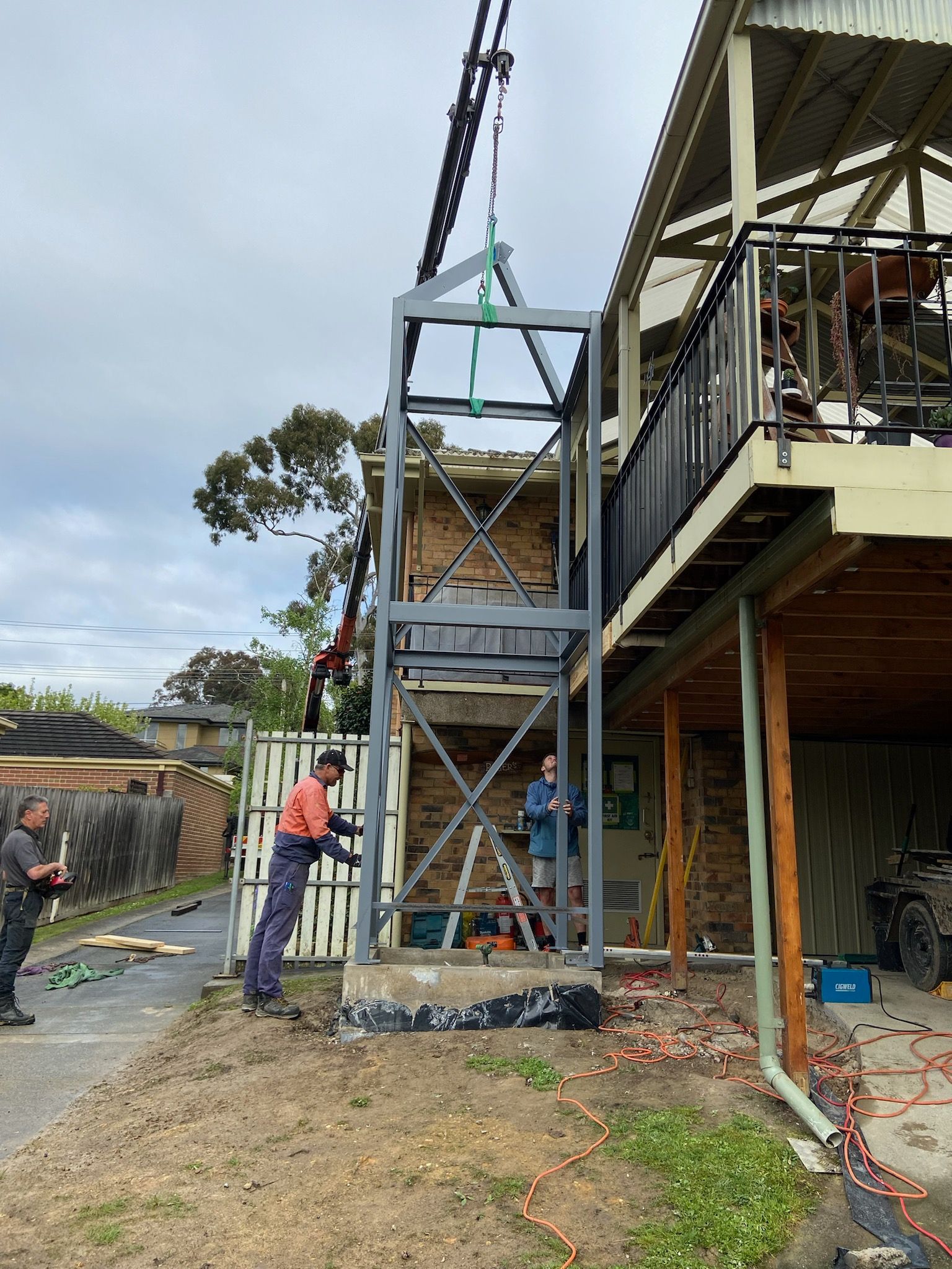 Construction workers installing a gray elevator shaft next to a house. Crane lifting it; workers in action.