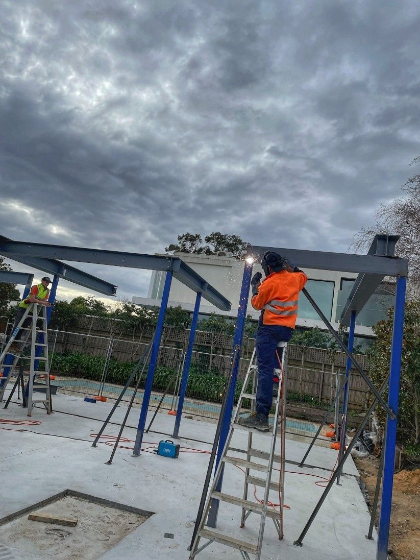 Construction worker welding steel beams on a cloudy day; another worker on a ladder nearby.