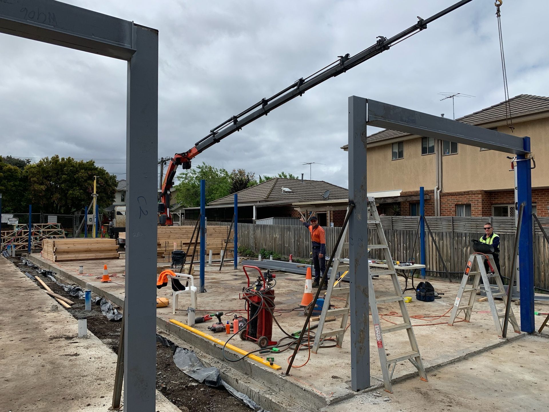 Construction site with crane lifting steel beams. Workers use ladders, with houses in background. Cloudy sky.