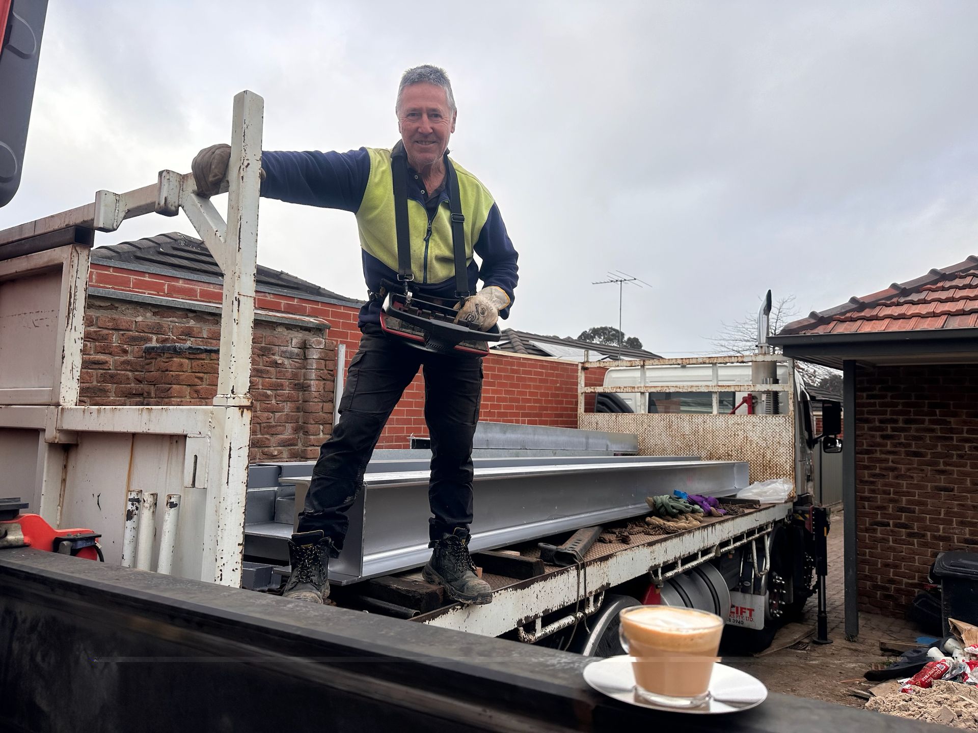 Construction worker on a truck, holding tools, with coffee. He's smiling. Grey sky, brick houses in the background.