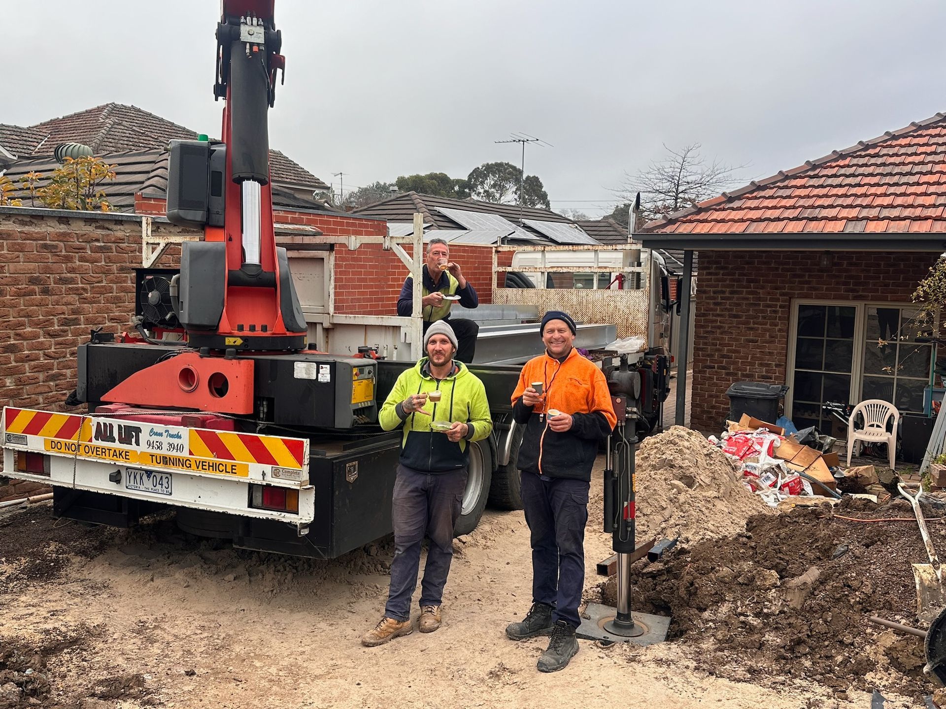 Three construction workers near a crane in a residential area; they hold drinks.
