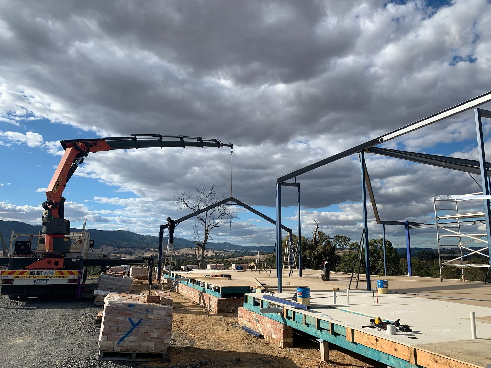 A crane lifting steel frame during construction. Cloudy sky with mountains in the background.