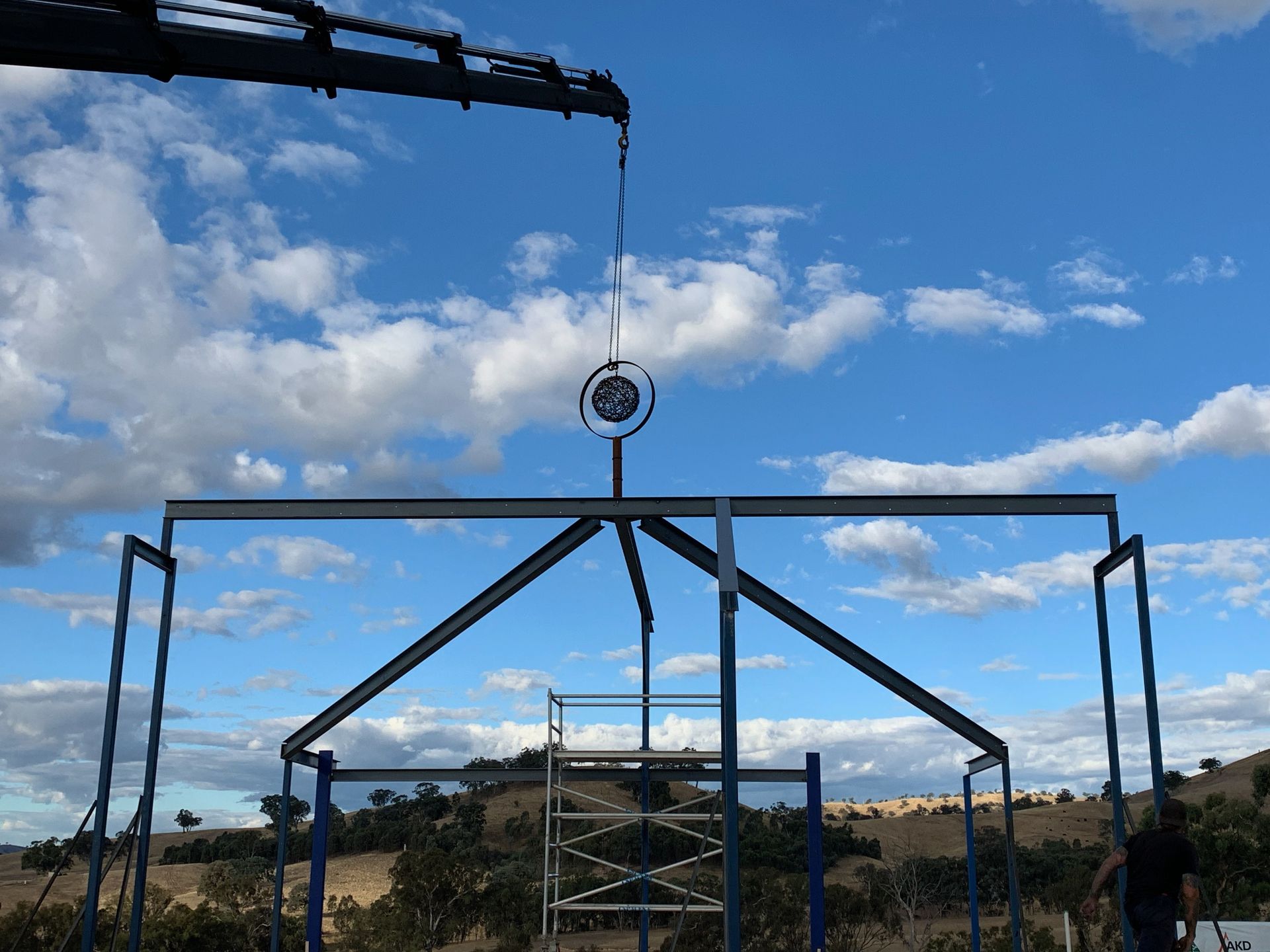Crane lowering a metal structure component against a blue sky. A man stands near the ground.