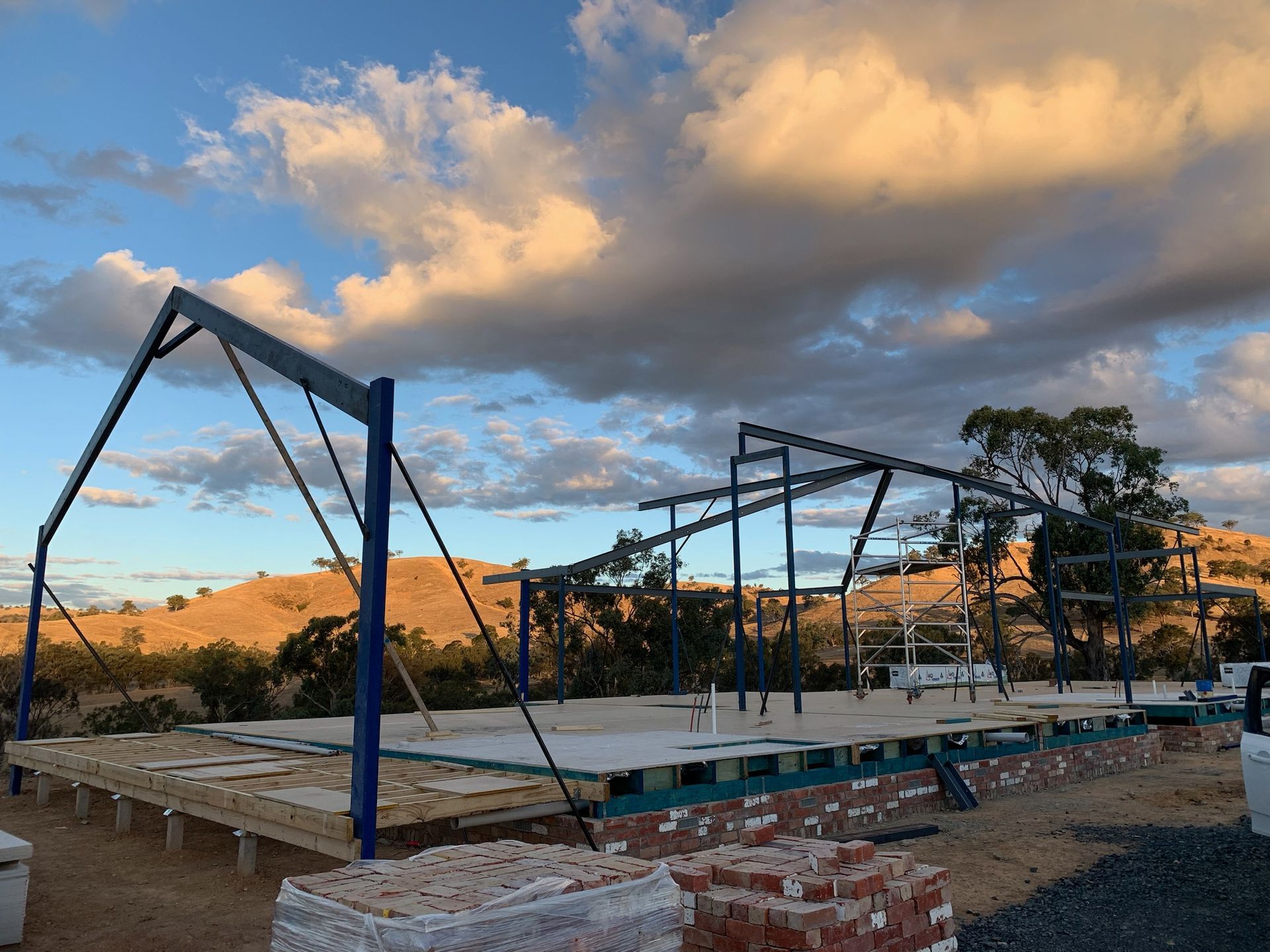 Construction site with blue steel frame, brick foundation, and wooden platform under a cloudy sky.