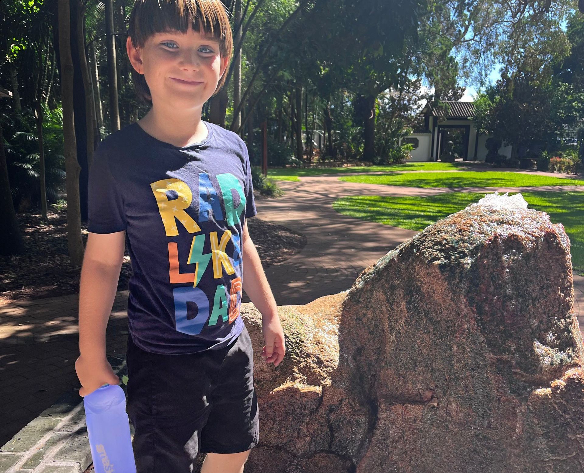 A boy standing outside next to a rock with his drink bottle - NDIS in Urangan, QLD