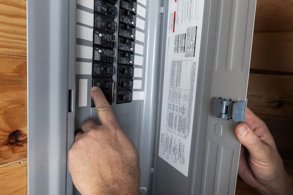 Hands turning on a breaker in an open electrical panel near a wooden wall.