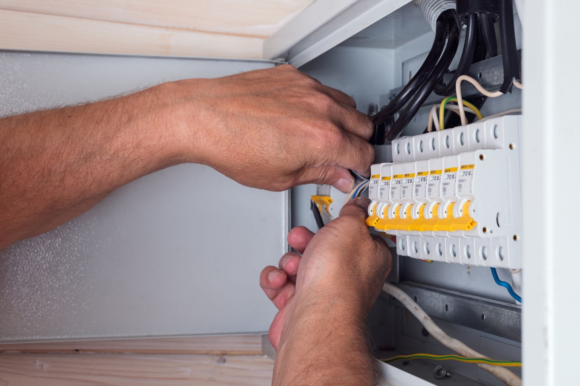 Hands wiring a white electrical breaker panel inside a cabinet.