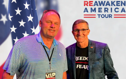 Two men posing at the Reawaken America Tour event backdrop, one wearing a VIP lanyard