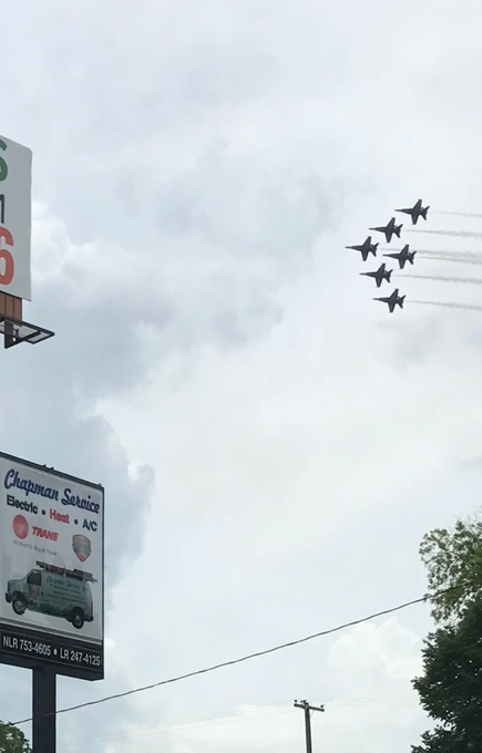 Six jets flying in formation across a cloudy sky above street signs and trees