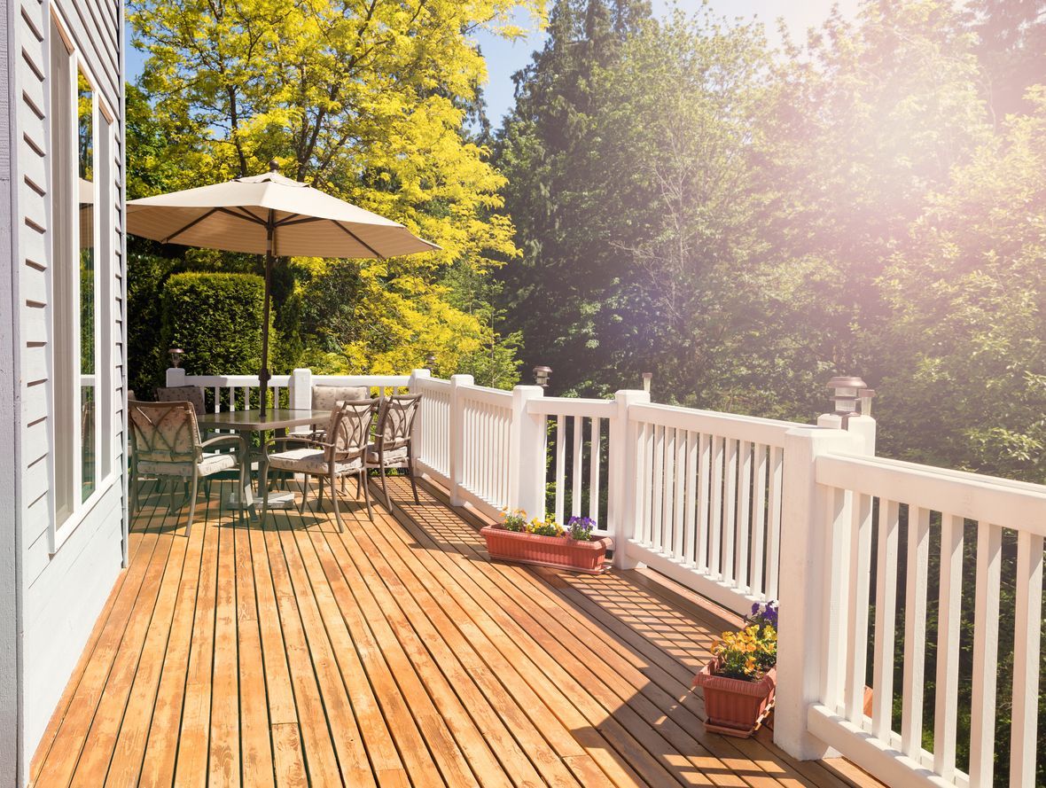 Sunlit wooden deck with white railing, patio table and umbrella, potted flowers, and trees beyond.