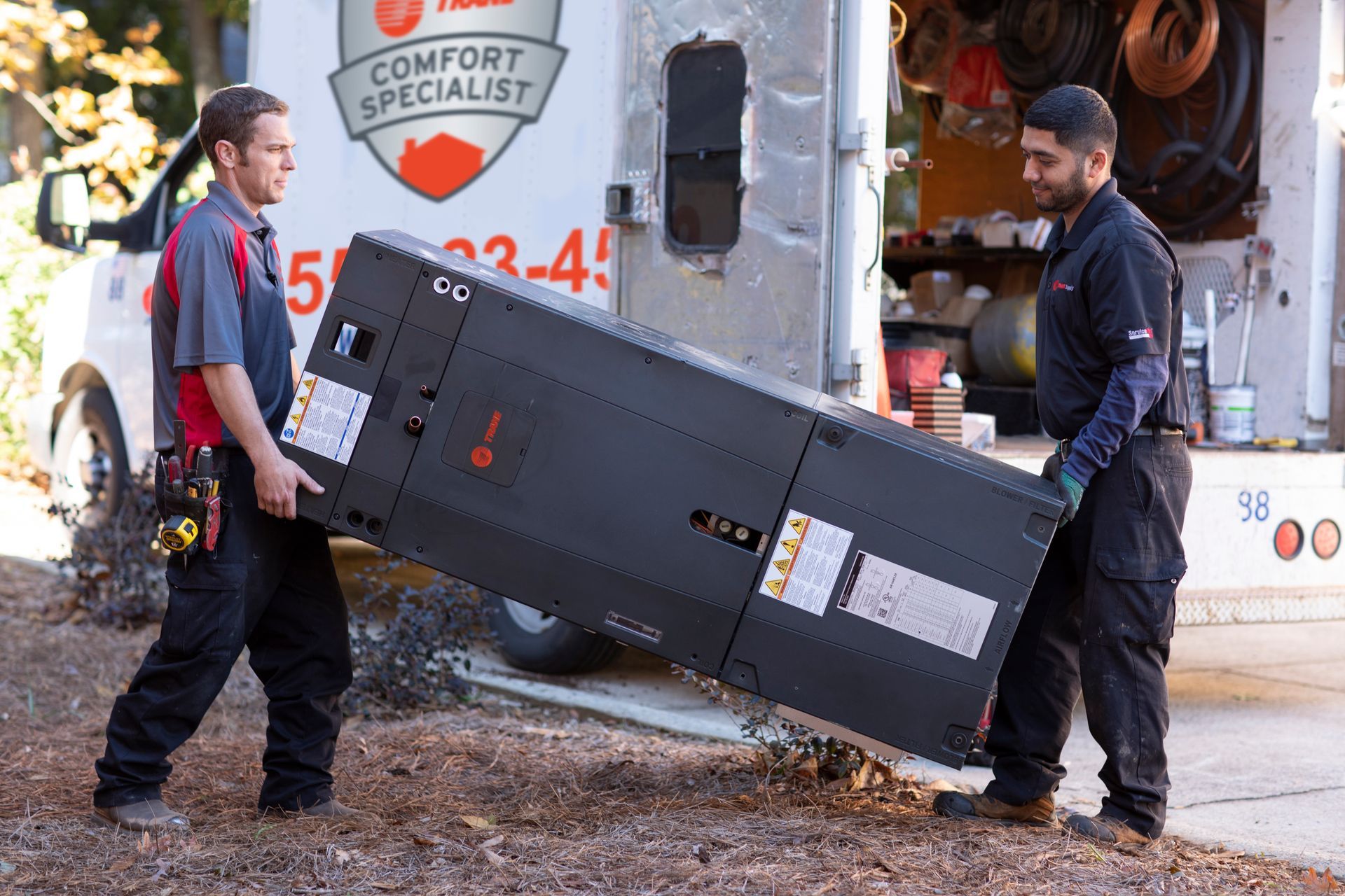 Two workers carry a large black air handler out of a van
