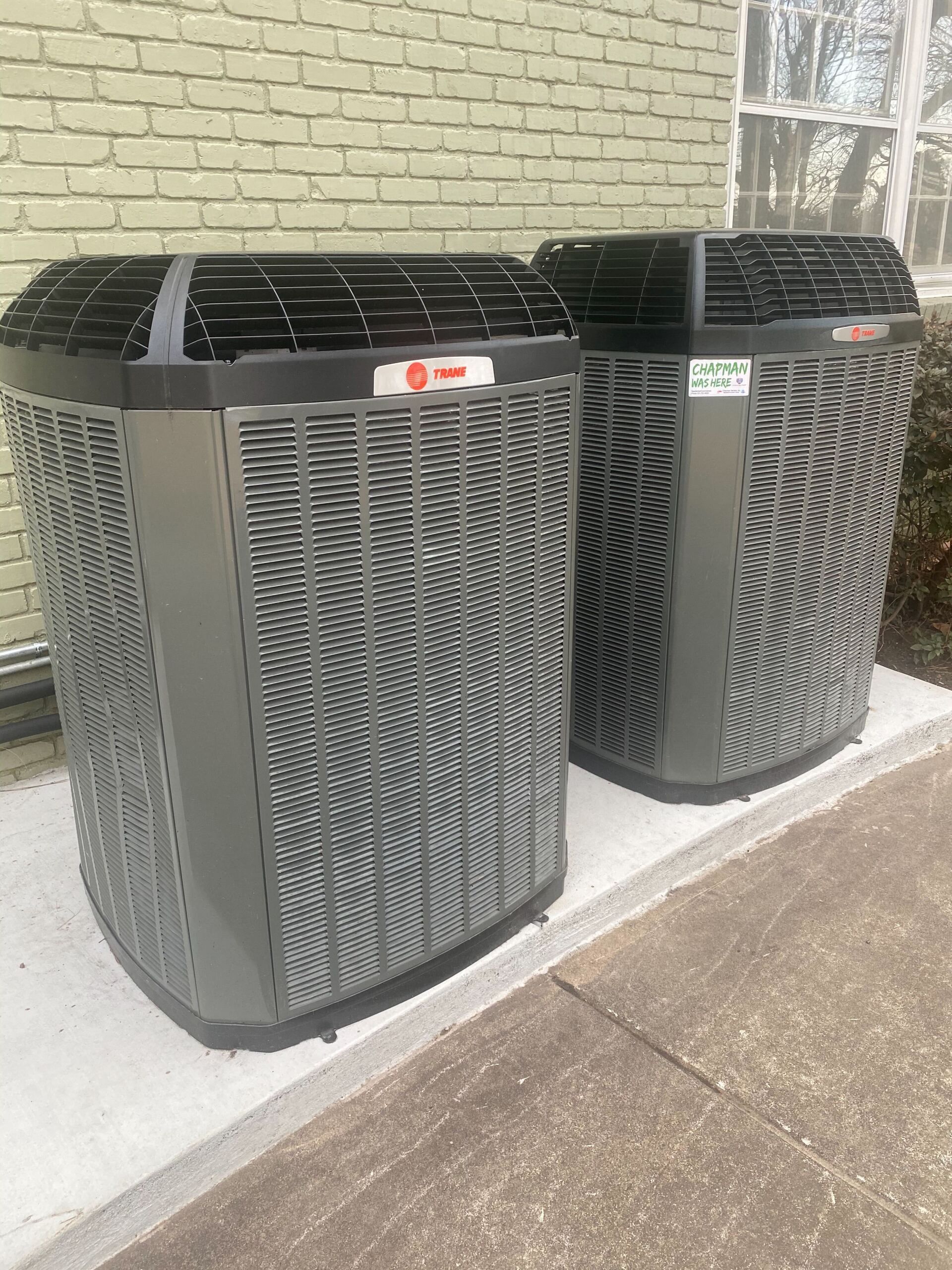 Two Trane outdoor air conditioner units beside a brick wall on a concrete sidewalk