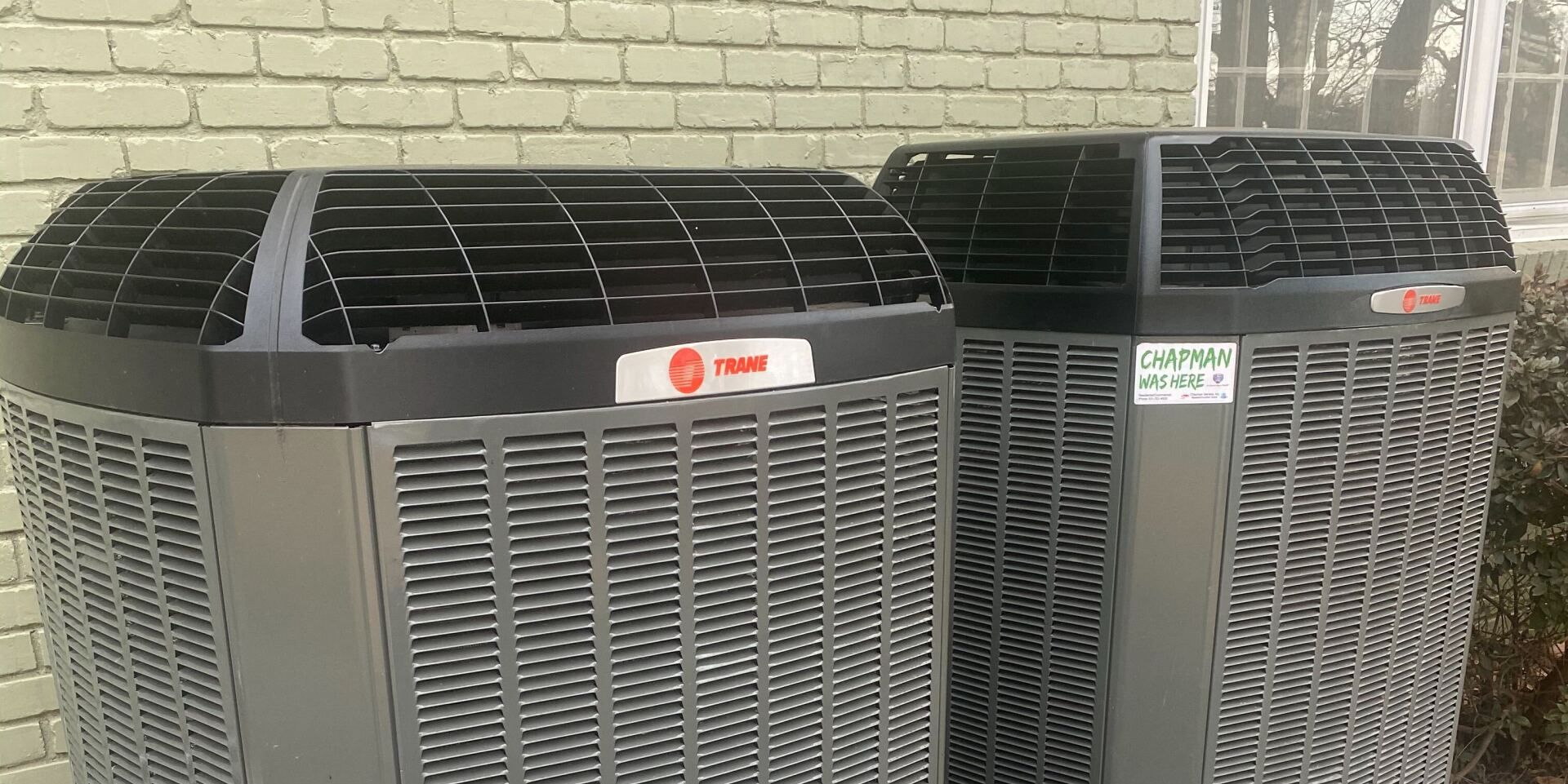 Two large gray HVAC units with black tops and labels, outdoors against a light brick wall.
