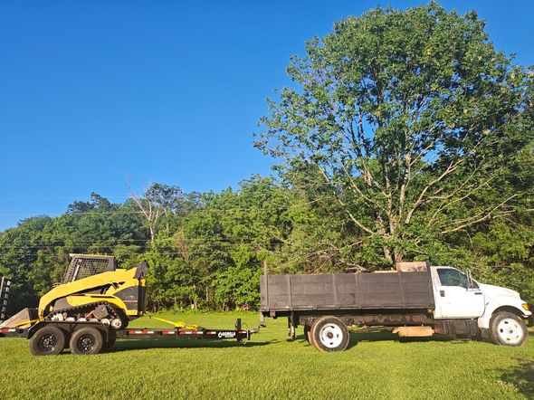 A yellow skid steer on a trailer is towed by a white dump truck on a grassy lawn under a blue sky.