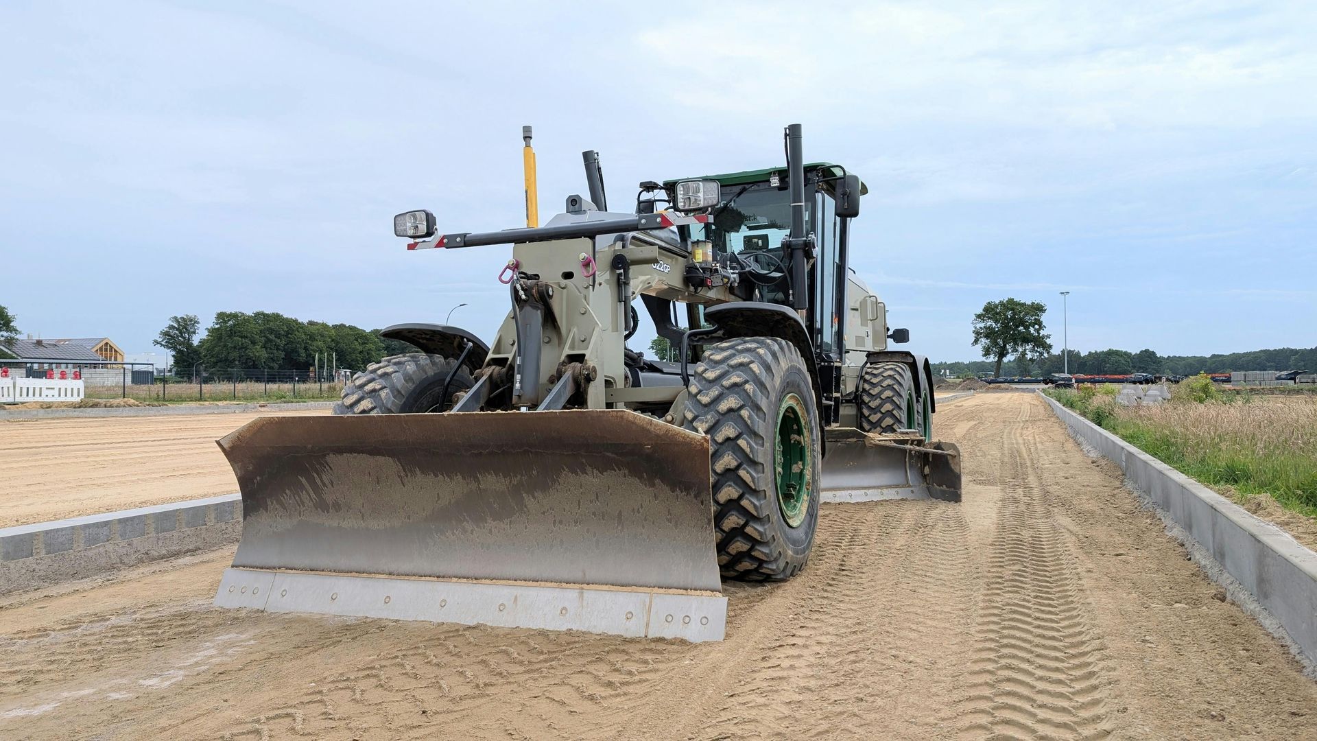 Motor grader leveling sand on a construction site with a cloudy sky in the background.