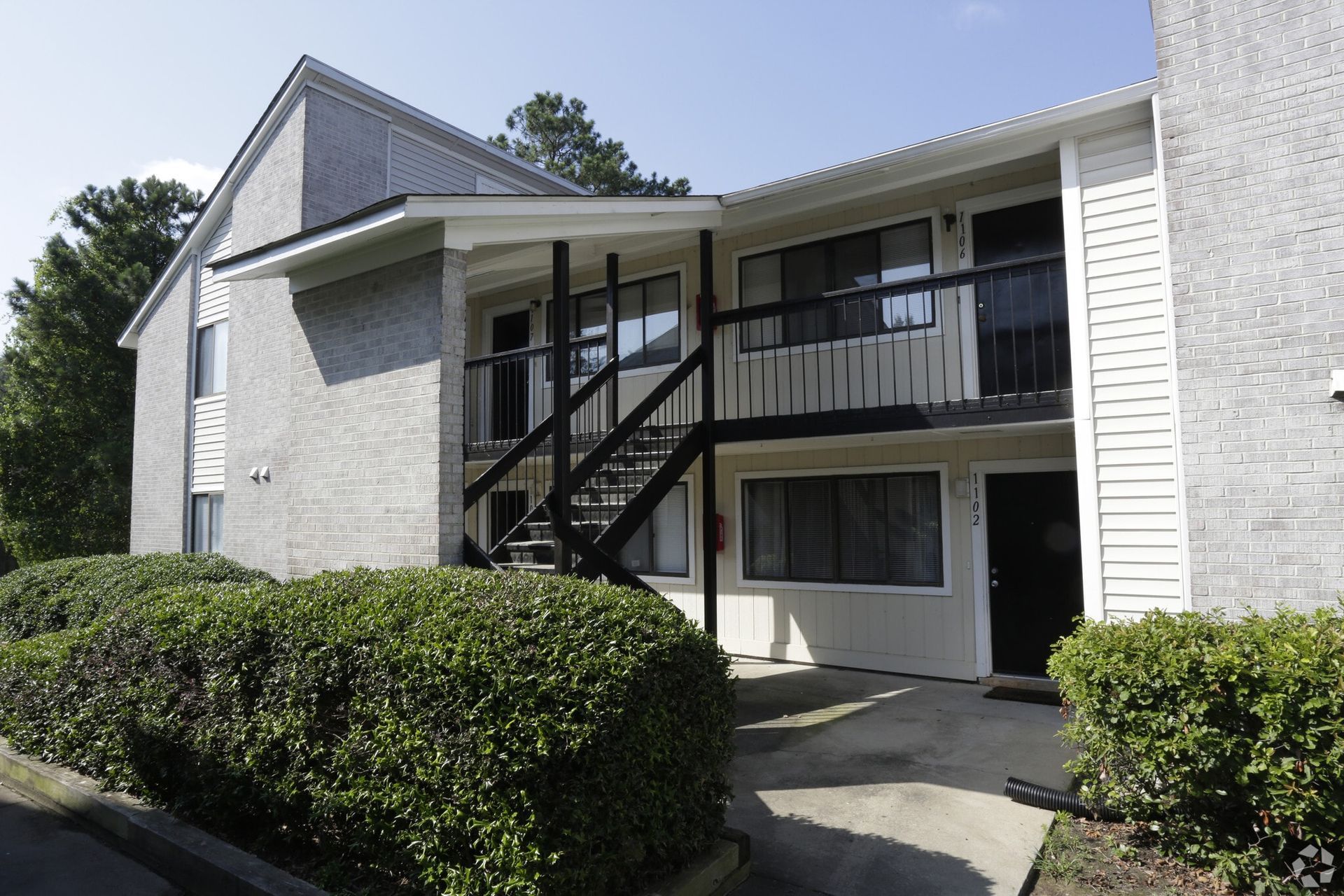 Two-story apartment building with brick exterior and black staircase. Bushes in front.