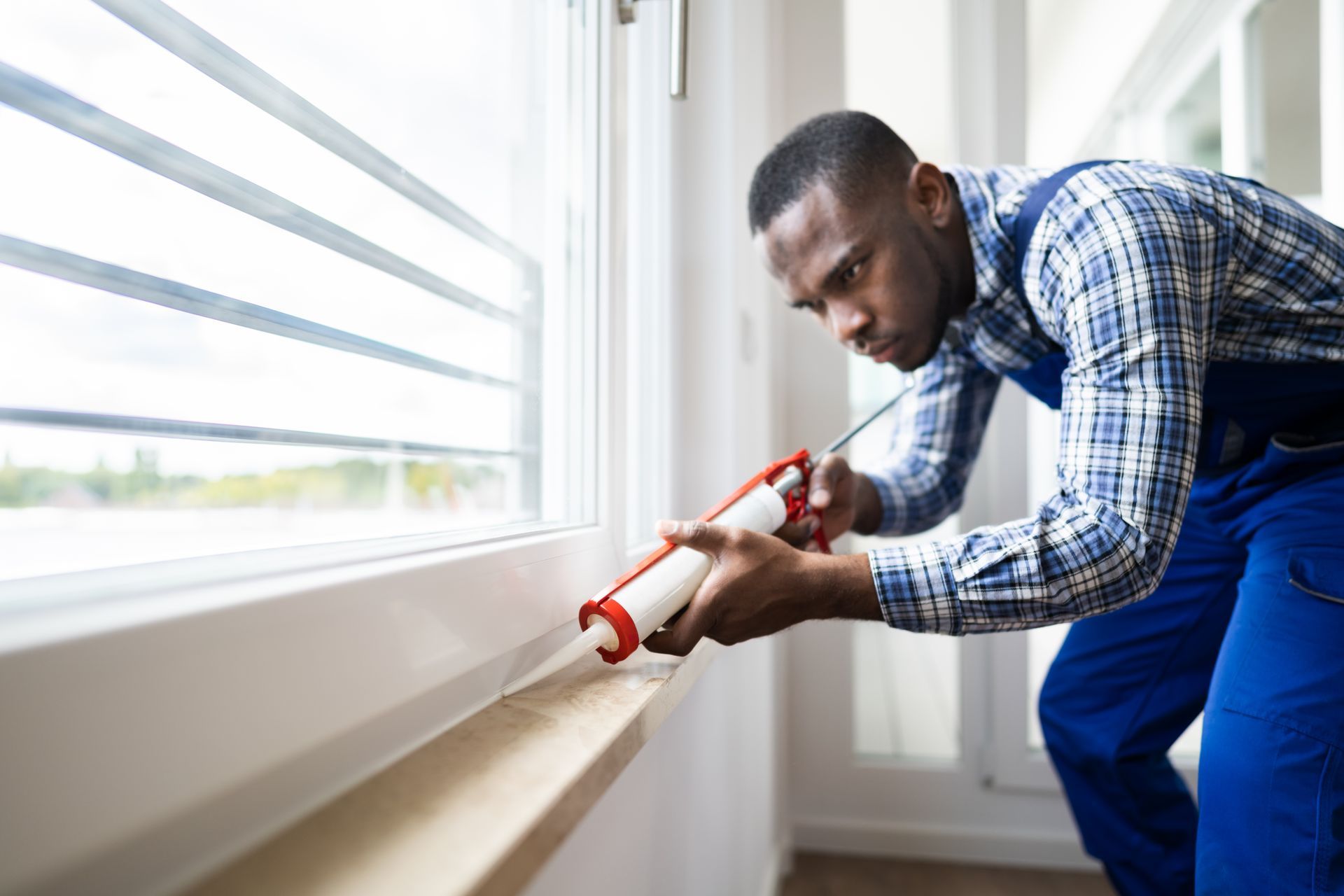 Man in blue overalls sealing a window frame with a caulk gun.