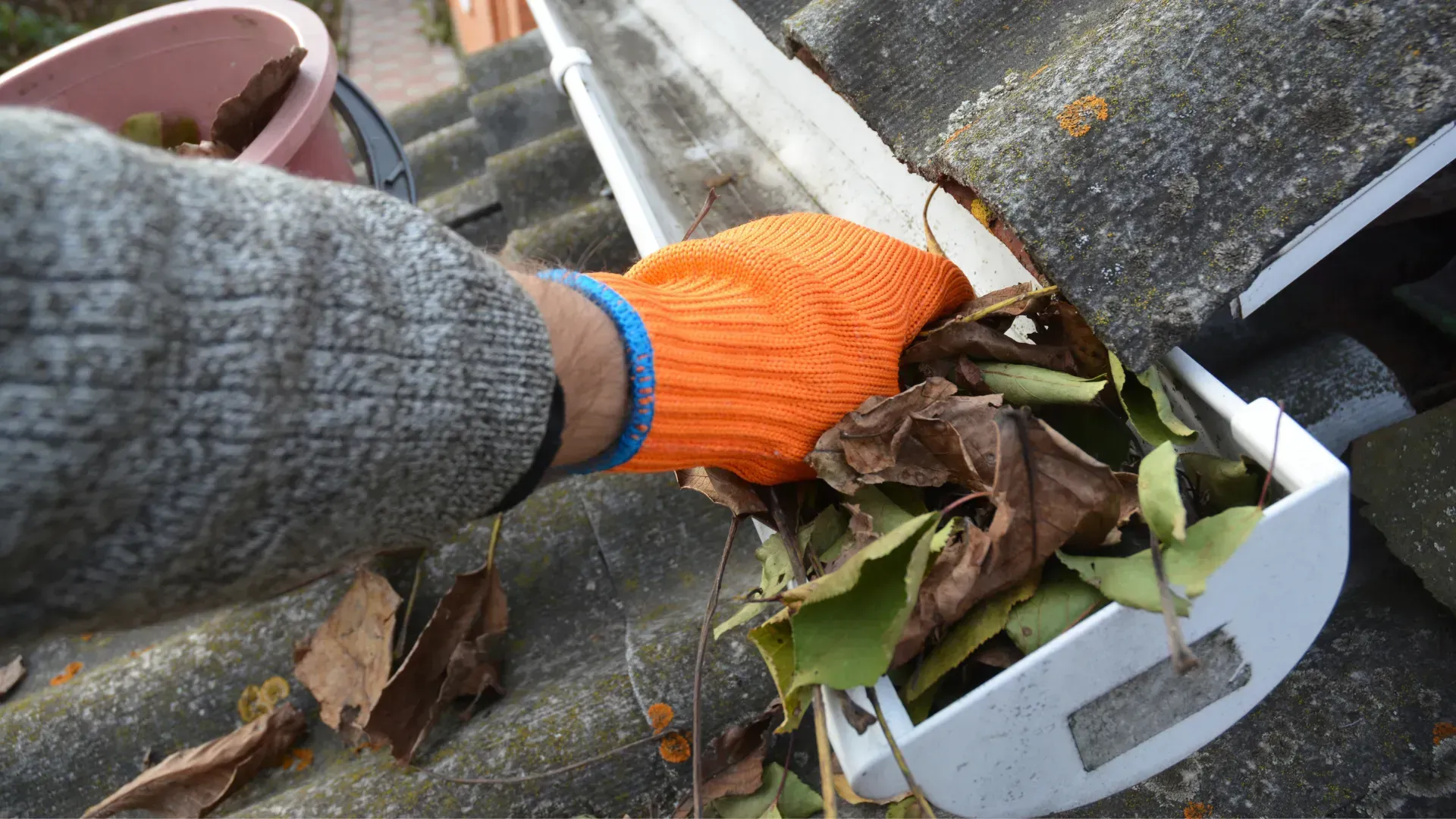 Person wearing orange glove, cleaning debris from a white gutter on a rooftop.