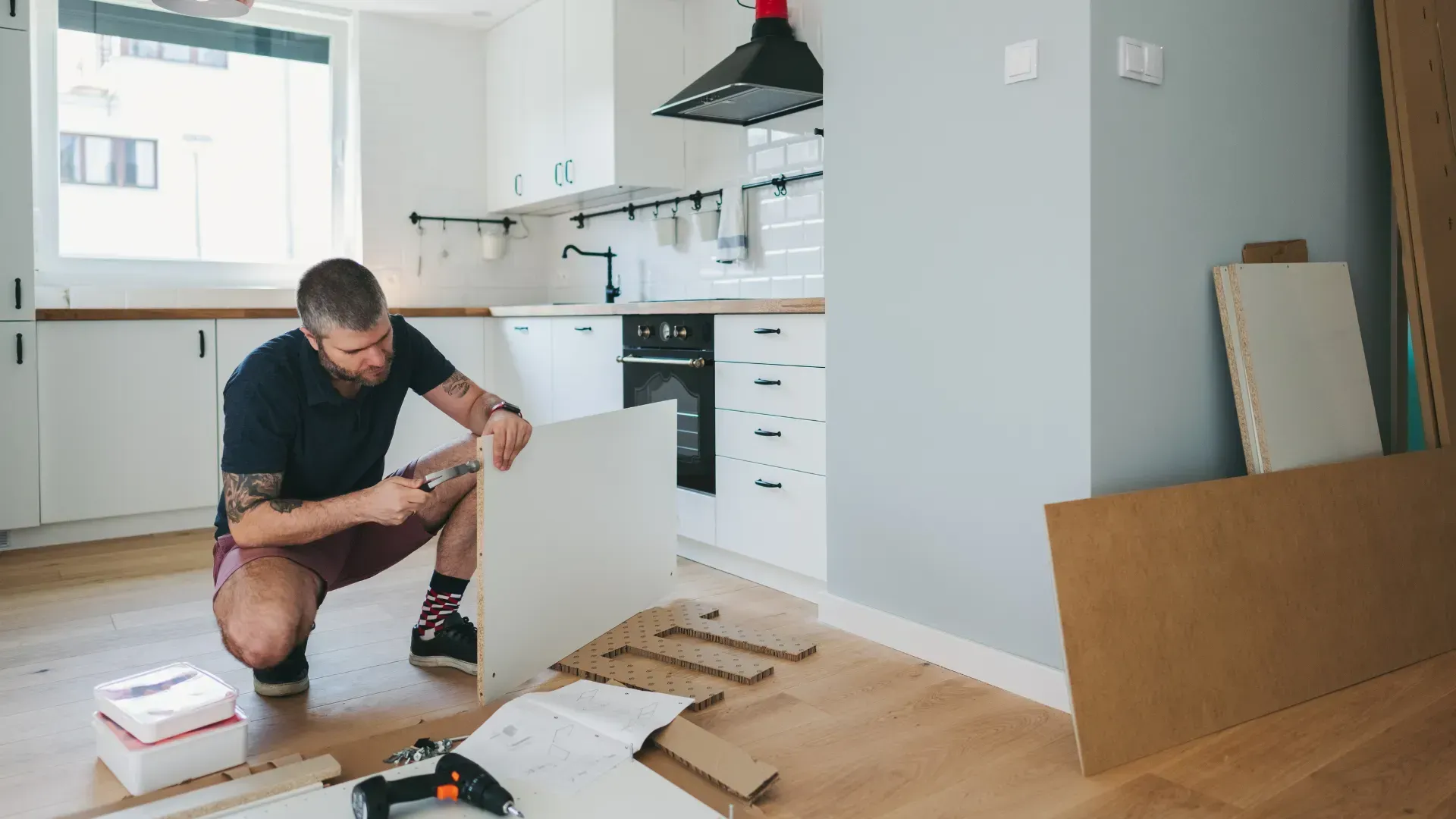Man assembling white kitchen cabinet in a light-filled kitchen, with instructions and tools scattered.