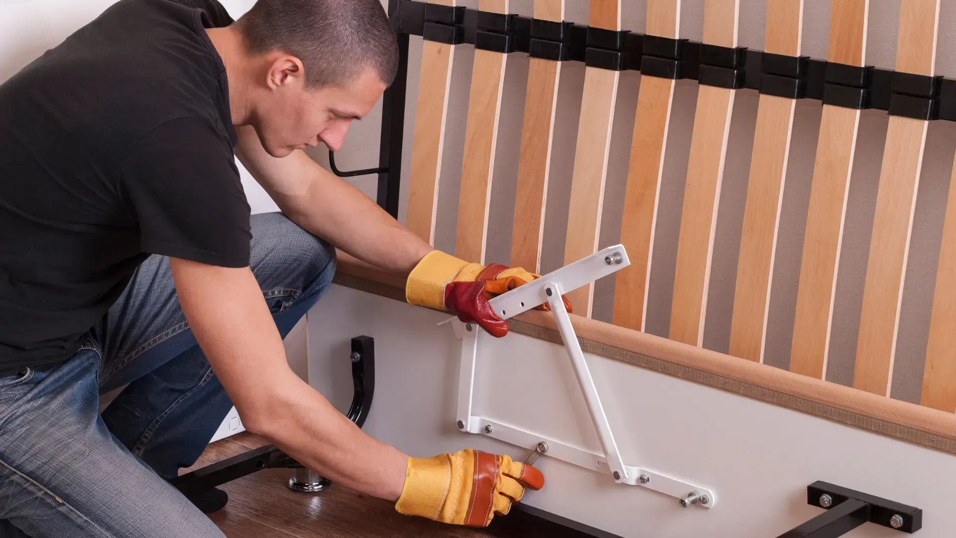 Man in work gloves assembling a bed frame, attaching a hinge with tools.