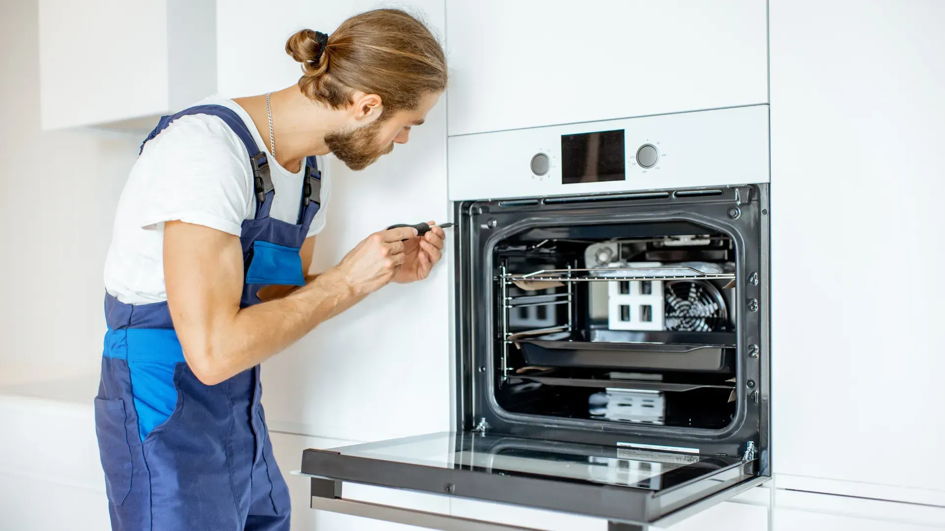 Man in blue overalls repairs an oven with a screwdriver in a white kitchen.