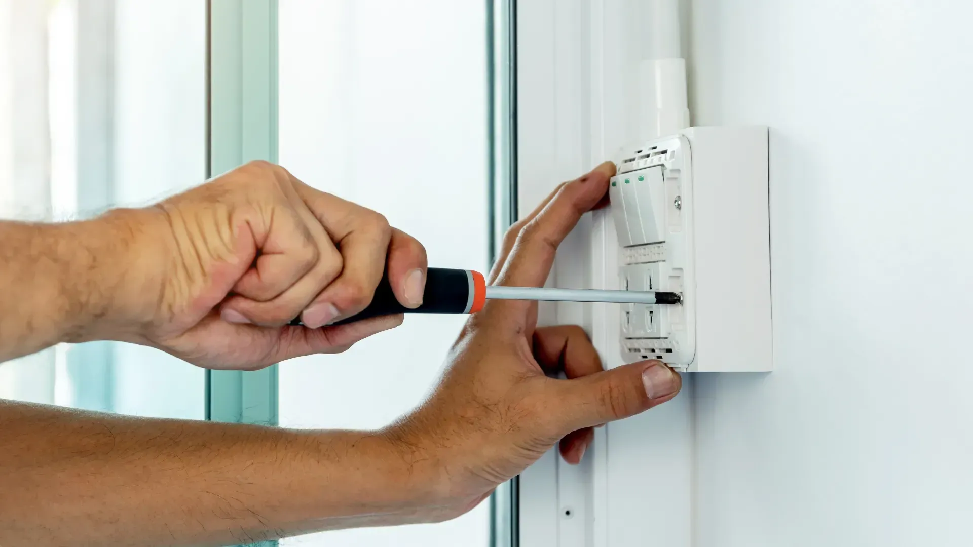 Person using a screwdriver to work on an electrical switch panel mounted on a white wall.