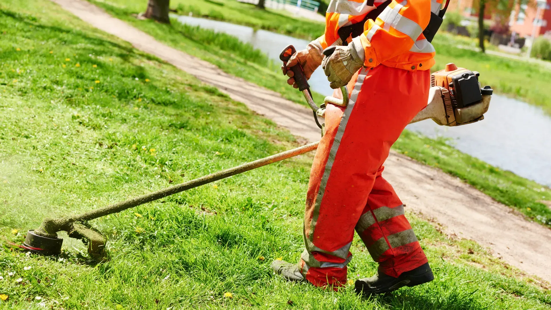 Person in orange work clothes using a weed wacker on a grassy embankment.