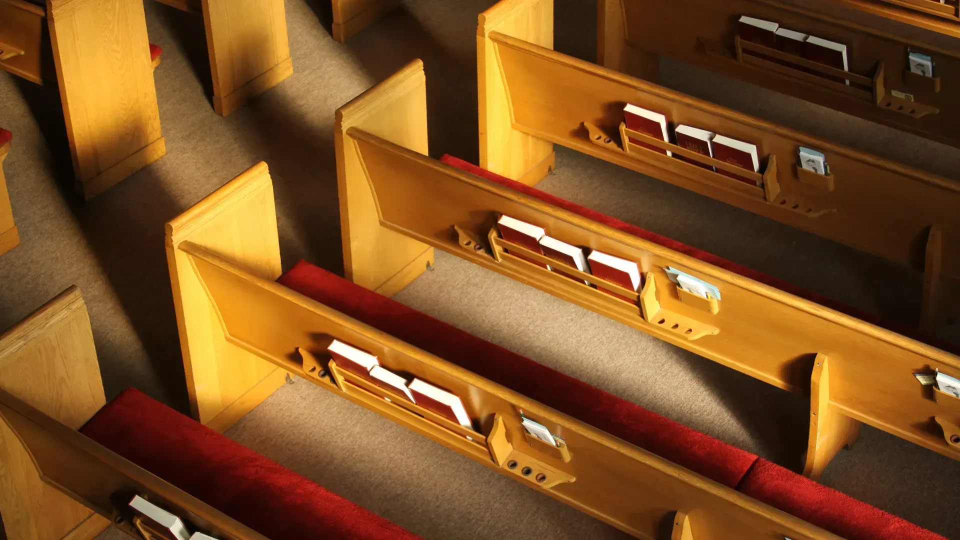 Rows of wooden church pews with red cushions. Sunlight casts shadows.