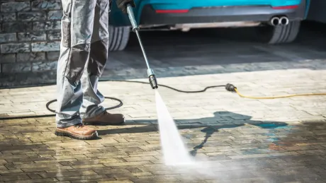 Person power washing a brick driveway in front of a teal car.
