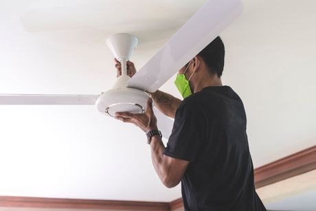 Man in black shirt and mask installing a white ceiling fan in a room.