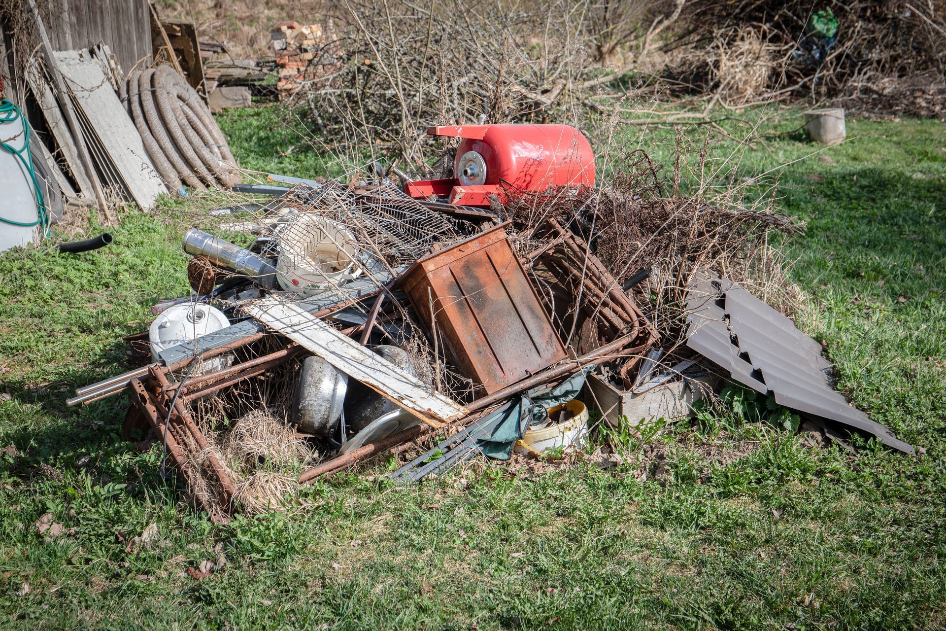 Pile of rusty metal and debris in grassy yard; red tank and wooden structure in the background.