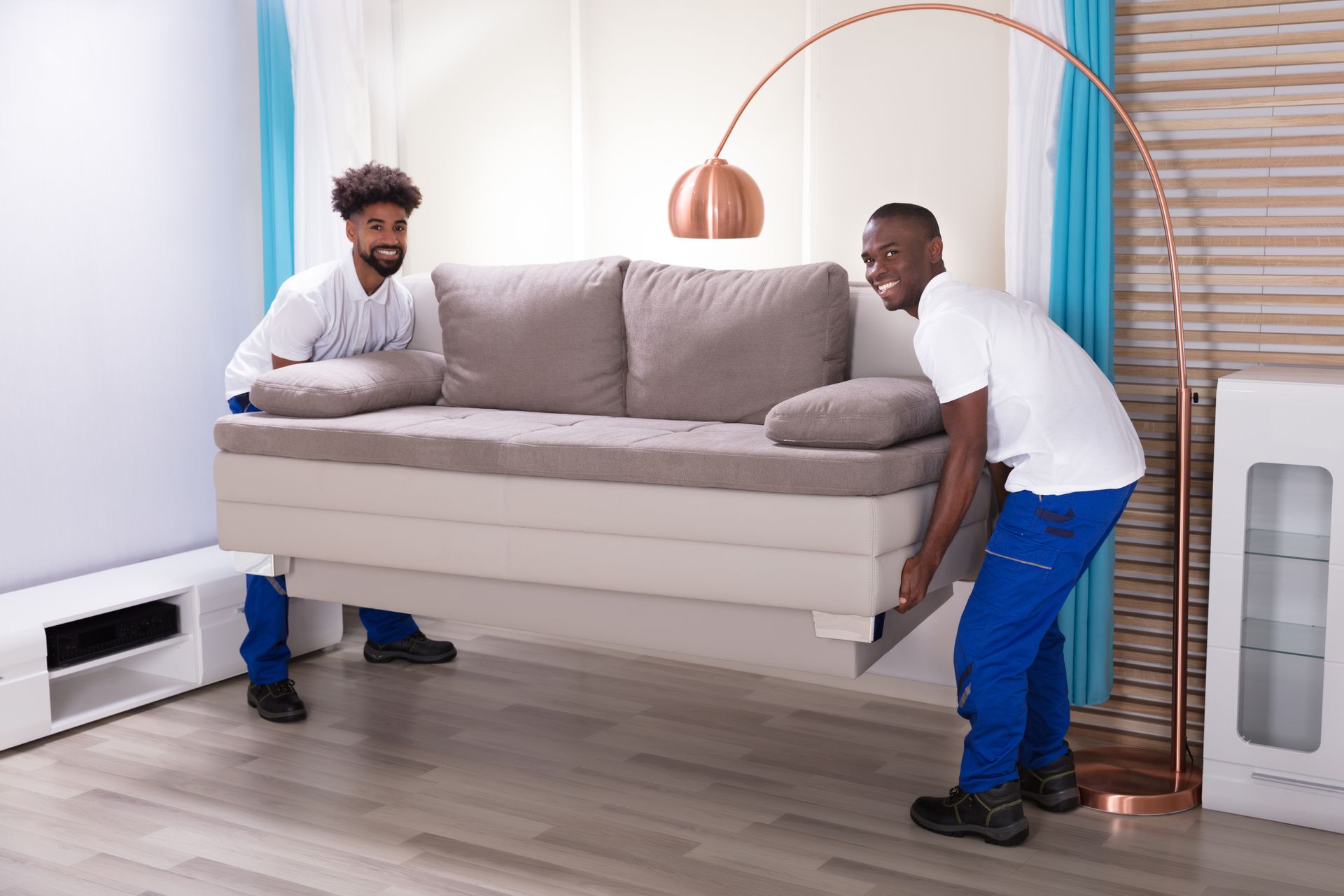 Two movers lift a beige couch inside a room, smiling. Blue pants, white shirts, under an arched lamp.