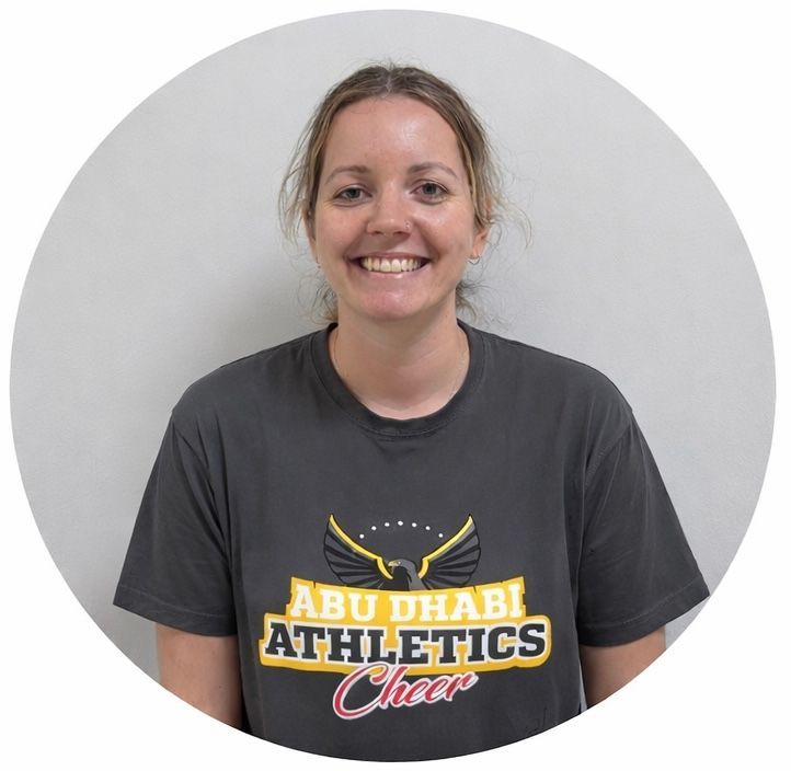 Woman in a gray Abu Dhabi Athletics cheer t-shirt smiles, against a white wall.