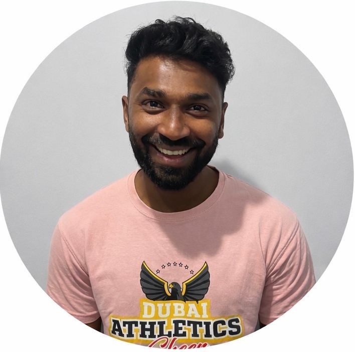 Man wearing pink t-shirt with Dubai Athletics logo, smiling in front of a gray wall.