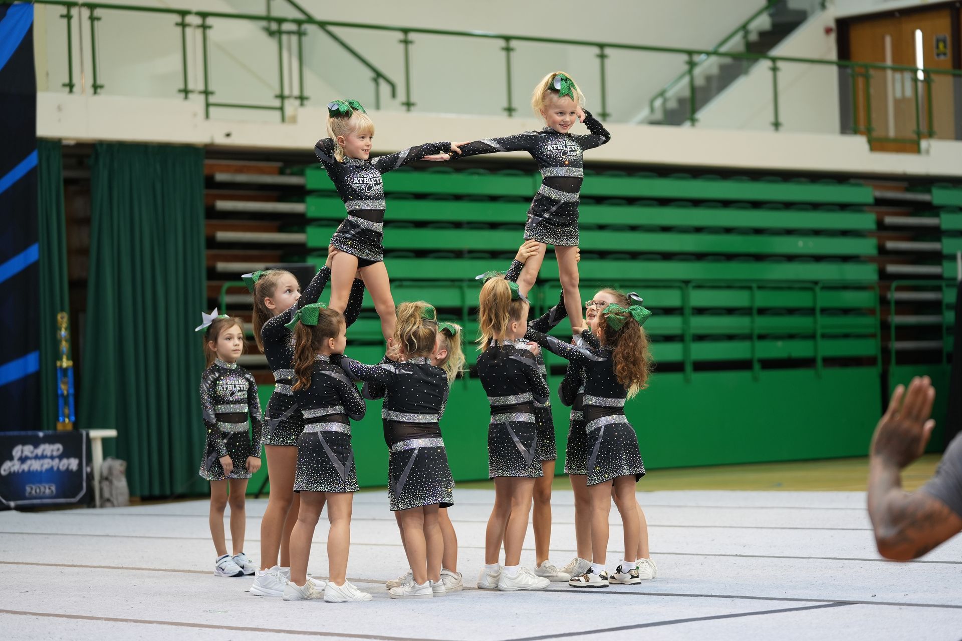 Cheerleaders performing a pyramid stunt indoors. Black and silver uniforms, green bows, and an audience in the background.