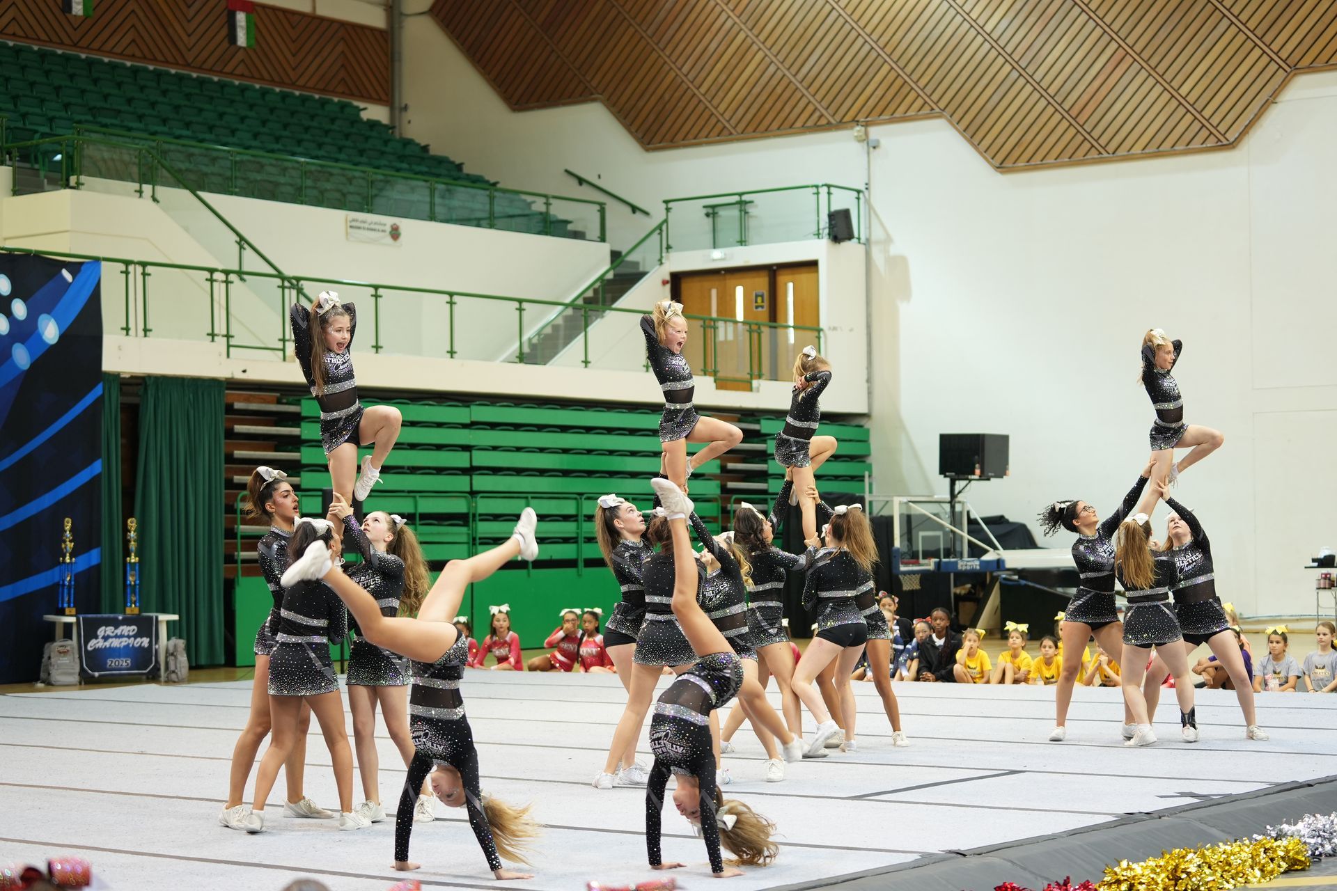 Cheerleaders performing stunts indoors: a pyramid and cartwheels. Black and silver uniforms.