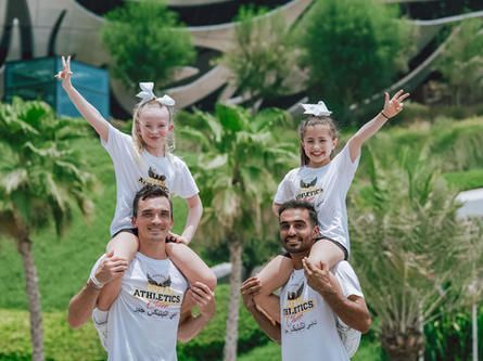 Two men carry two girls on their shoulders in front of a tropical setting; all are smiling and wearing matching shirts.