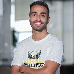 Man in a white t-shirt smiles with arms crossed; the shirt has a logo, blurred background.