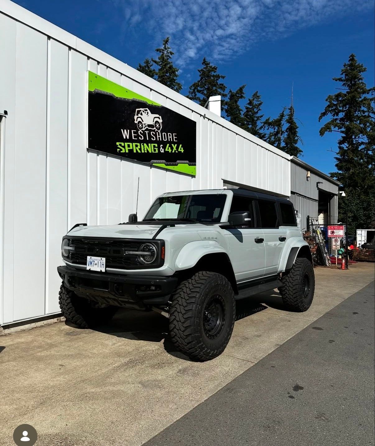 White Ford Bronco with large tires in front of a shop; Westshore Spring 4x4 sign. Blue sky.