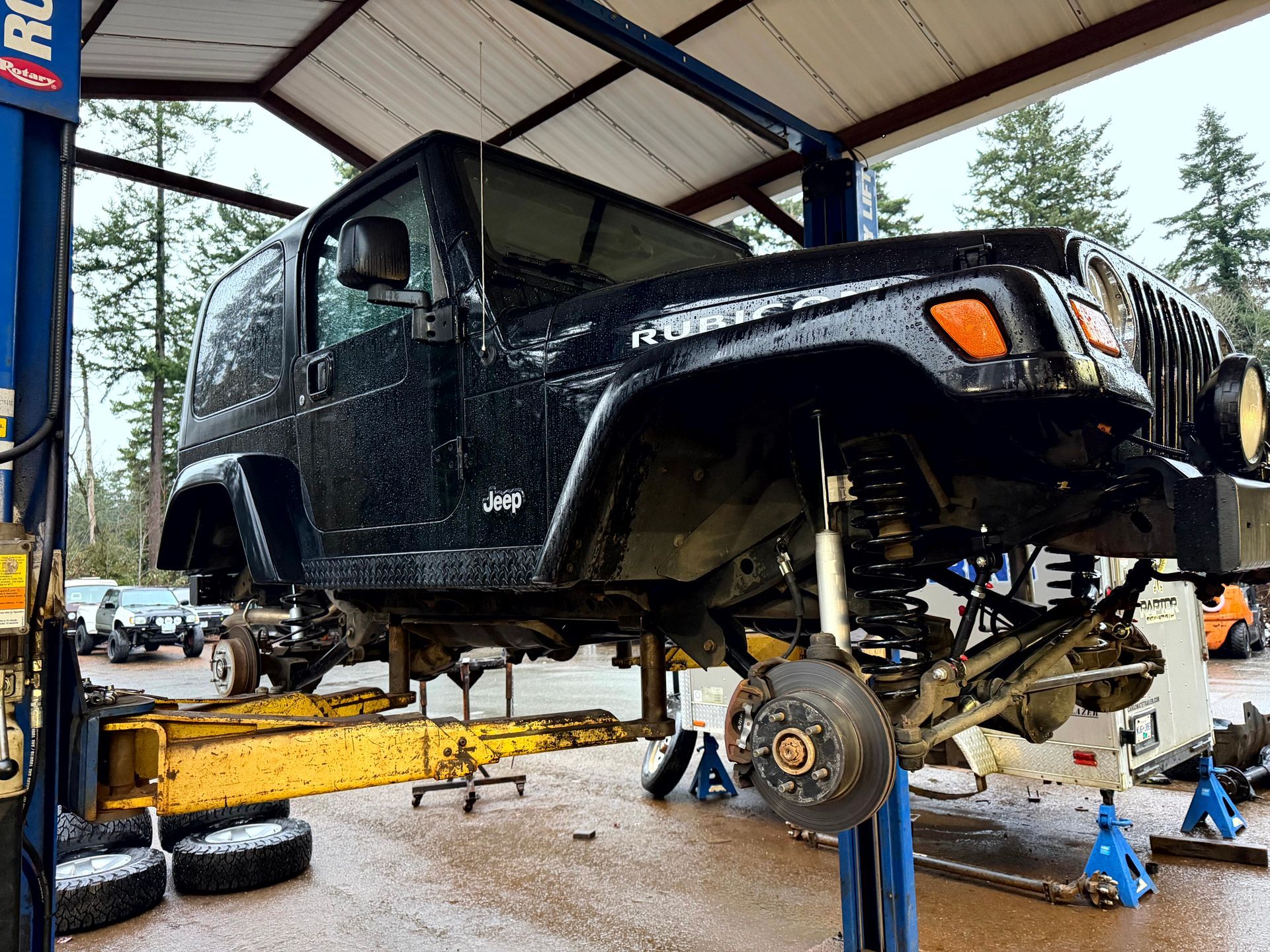 Black Jeep Wrangler on a lift in a repair shop, undergoing work; wheels and suspension are removed.