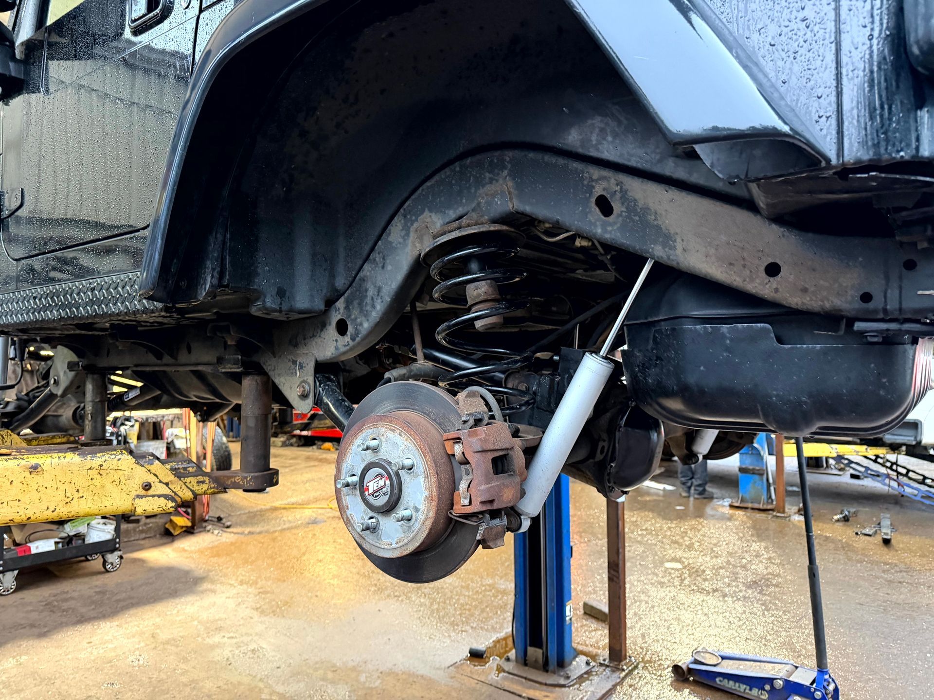 Black Jeep rear wheel raised, revealing suspension, brake system, and shock absorber in a repair shop setting.