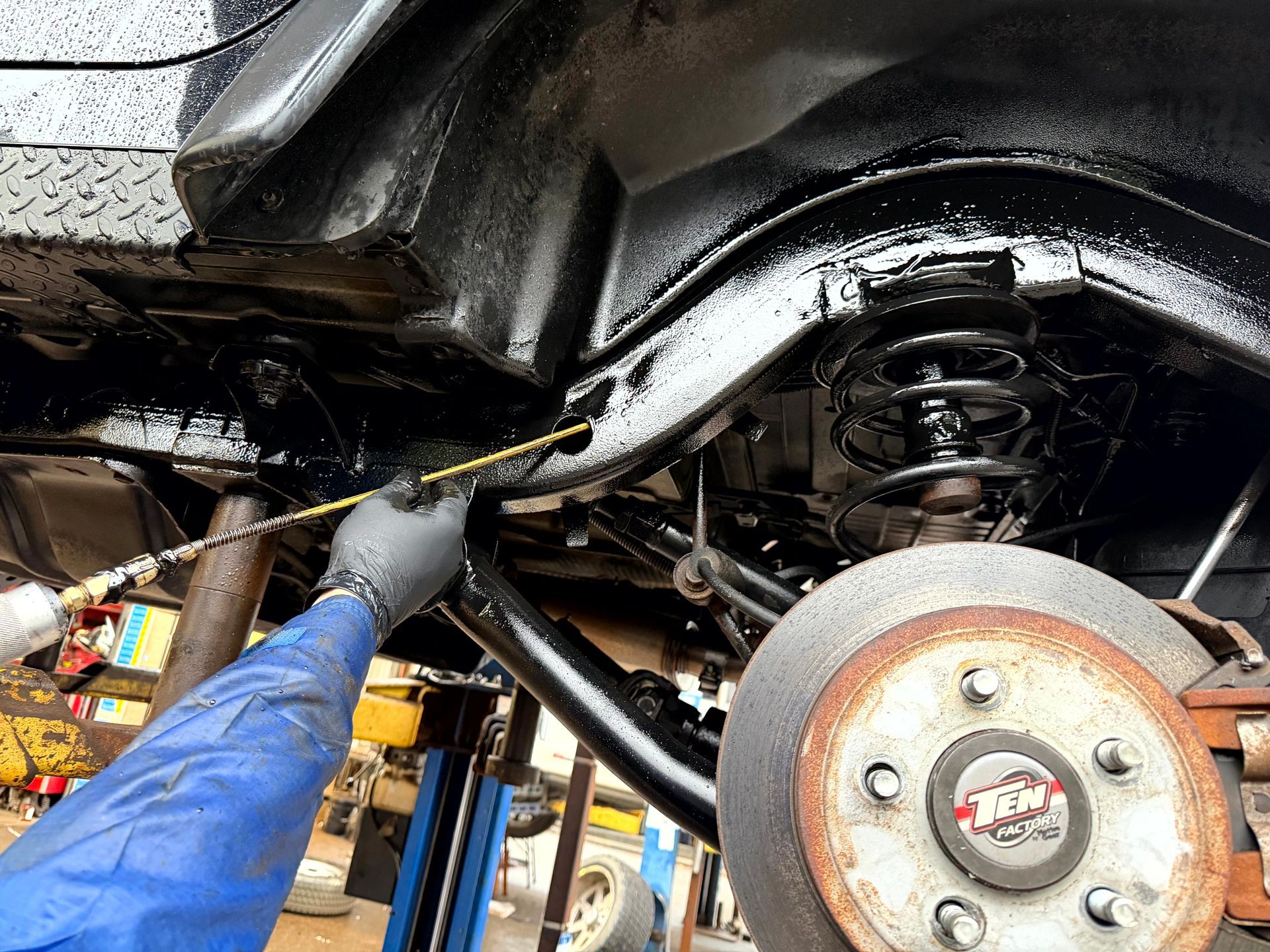 A person sprays black undercoating on a vehicle's underside, near a tire and suspension, indoors.