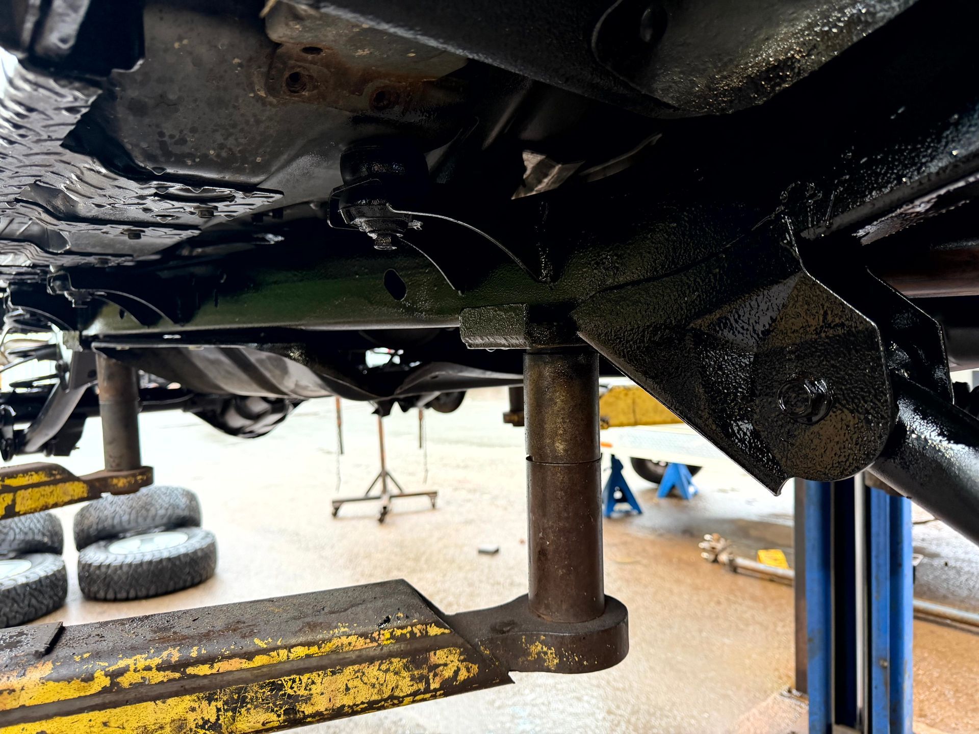 Undercarriage of a vehicle raised on a lift; black painted metal, supported by a jack stand in a shop.