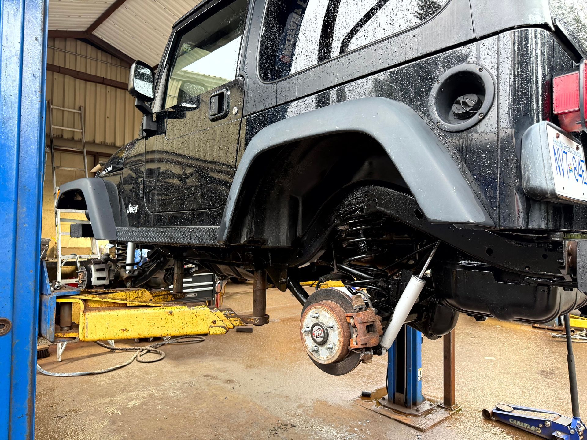 Black Jeep Wrangler on a hydraulic lift in a garage, rear wheel removed.