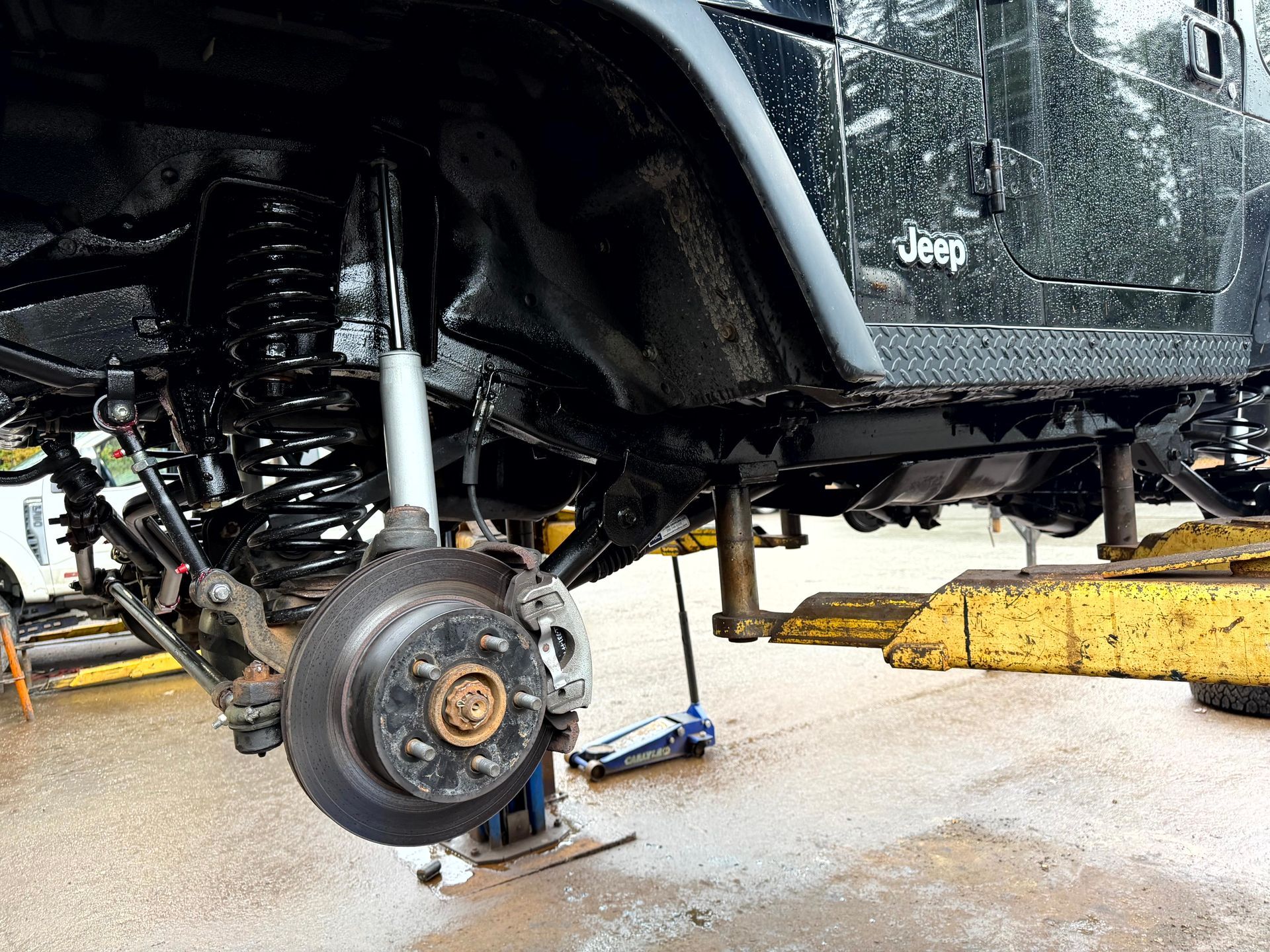 Black Jeep undercarriage on a lift, brake rotor exposed. Suspension components visible, ground is wet.