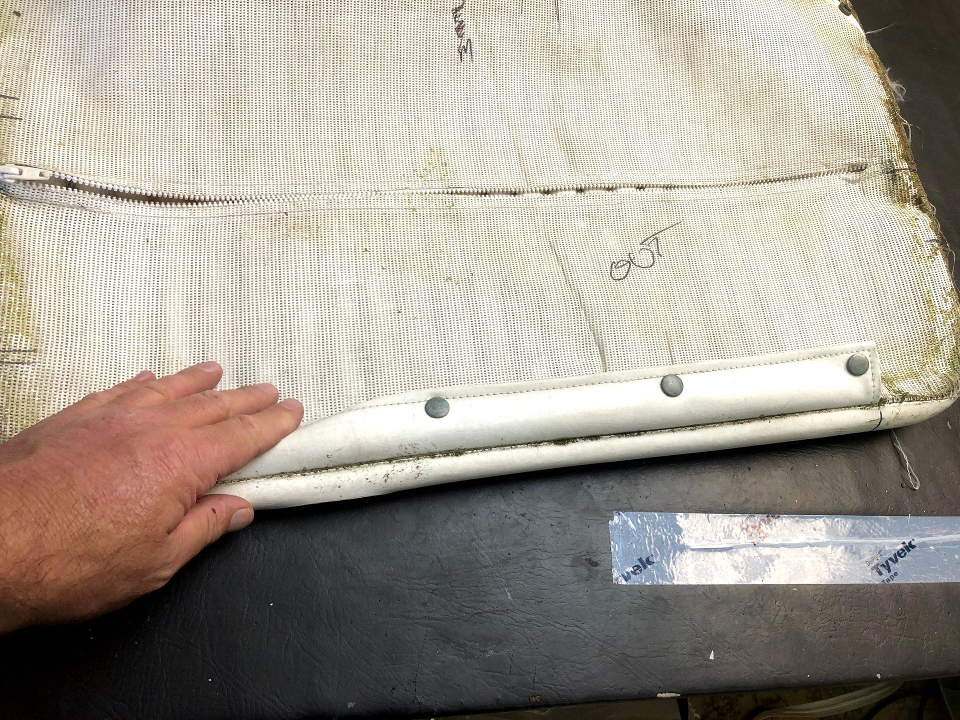 Hand holding the edge of a white mesh fabric piece with metal rivets, likely for furniture repair on a dark surface.