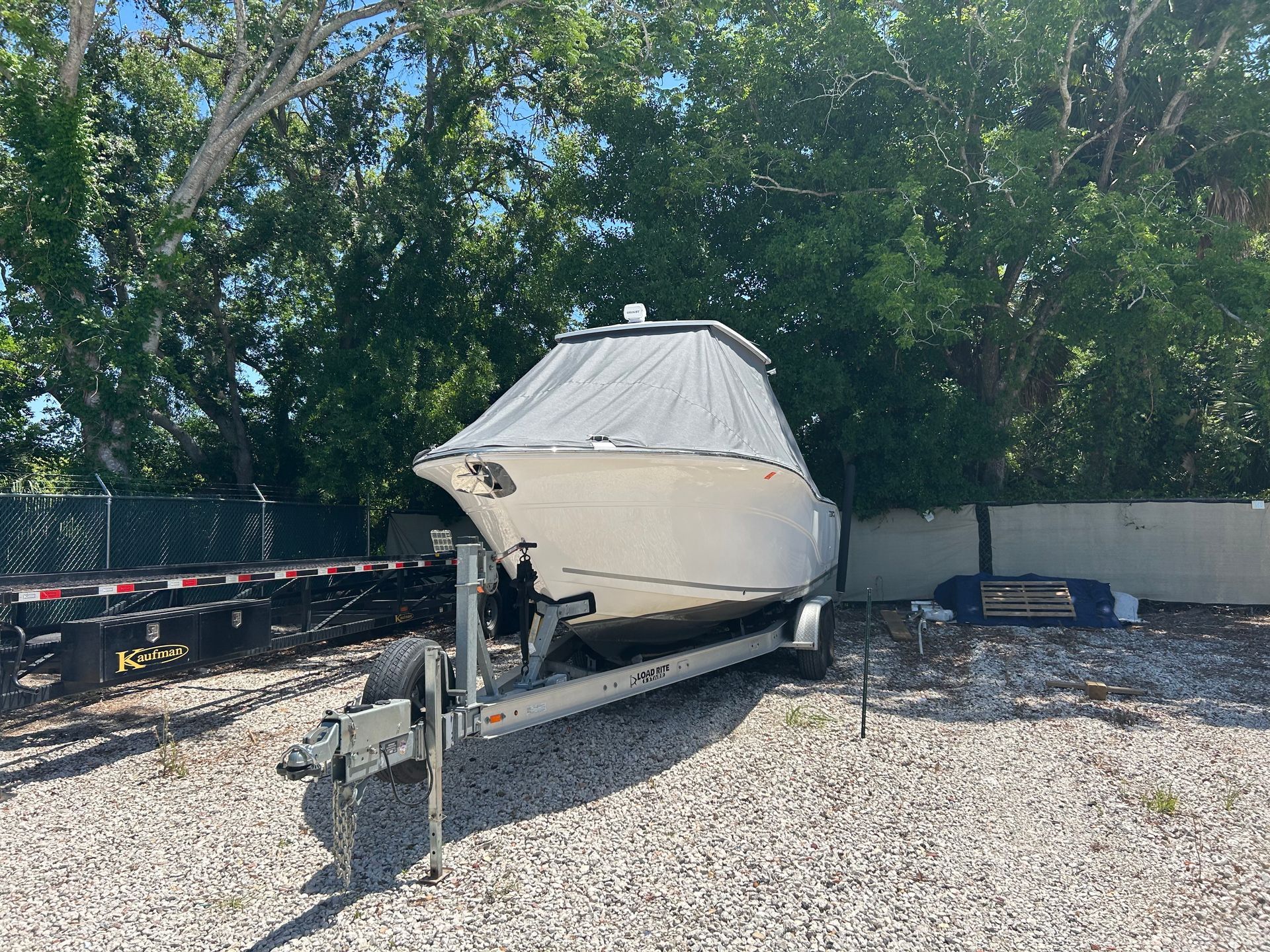Boat on a trailer, beige hull, covered cabin, parked on gravel, surrounded by trees.