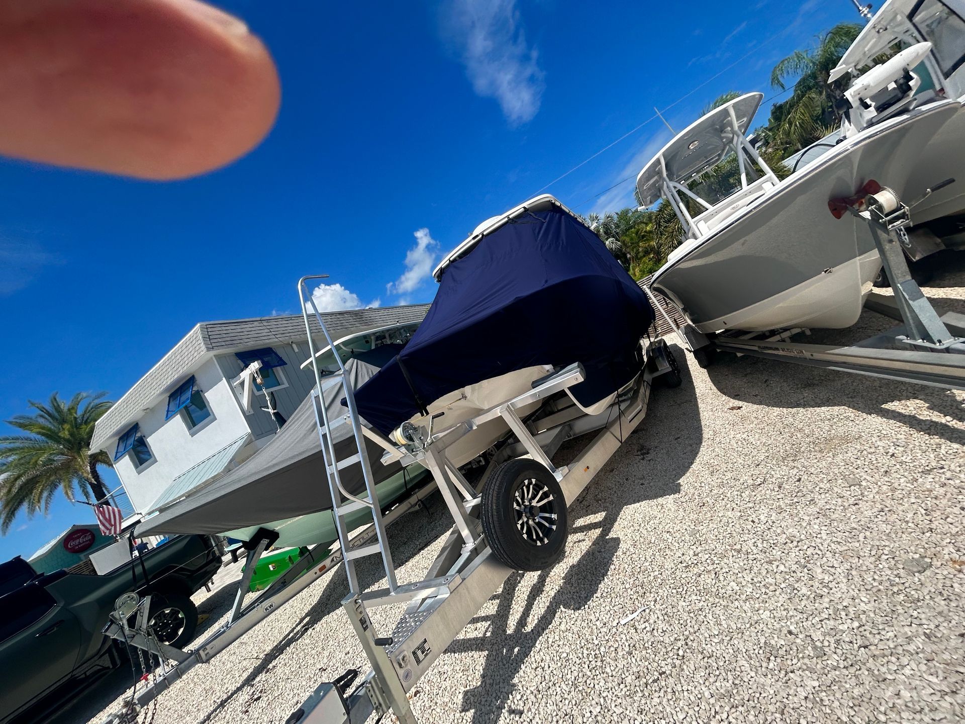 Boats on trailers under a bright blue sky. One boat covered by blue fabric. Setting: outdoors.