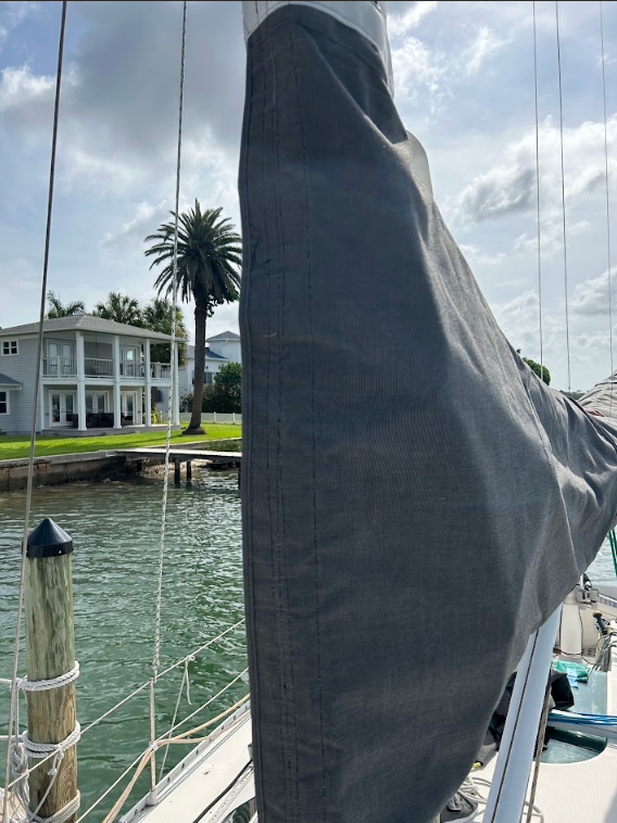 A sailboat's rolled-up gray sail on a sunny day with a house and palm tree in the background.
